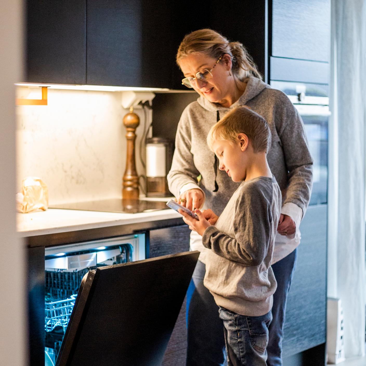 Mãe ensinando o filho a usar o aplicativo móvel enquanto opera a máquina de lavar louça na cozinha de casa