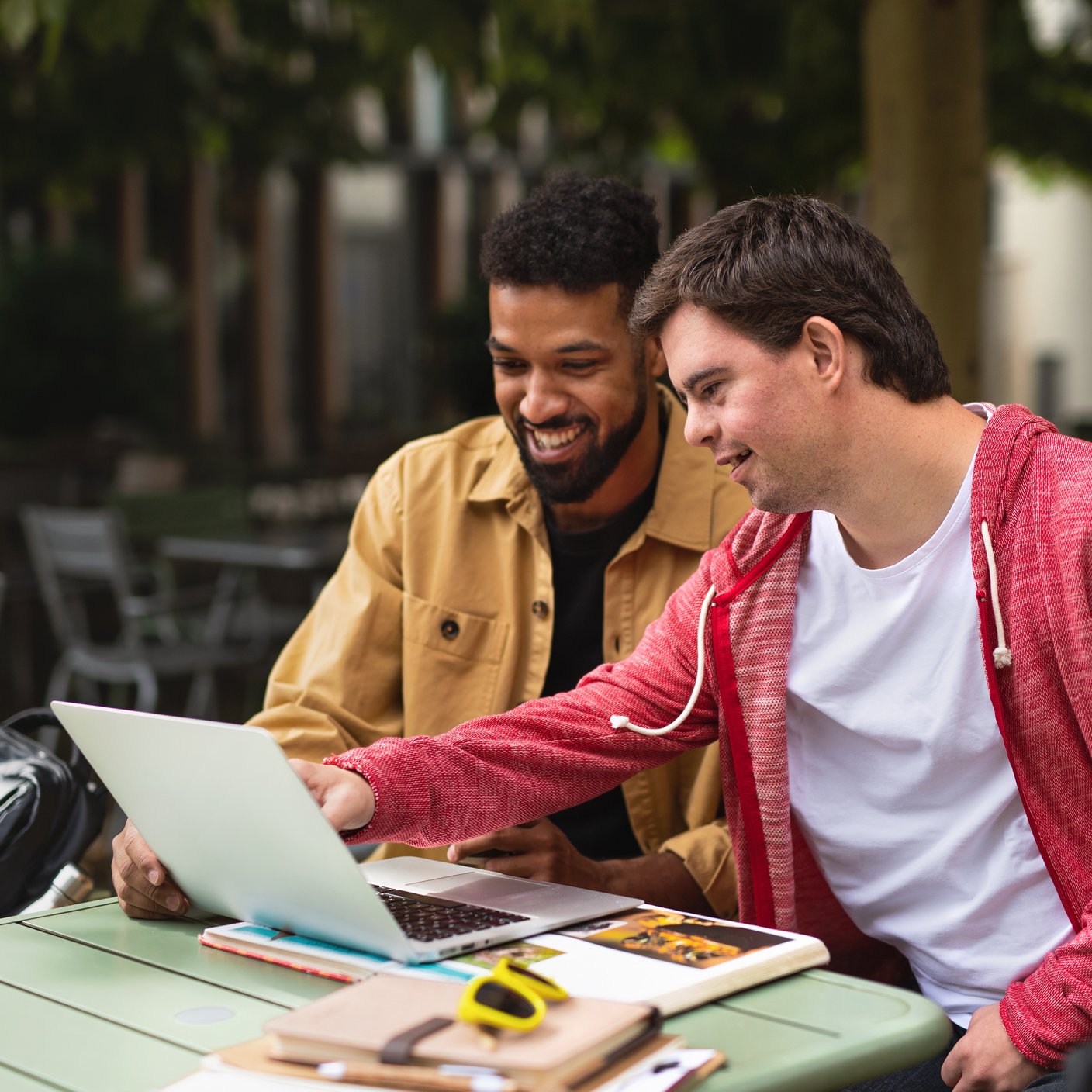 A young man with Down syndrome with his mentoring friend sitting outdoors in cafe using laptop.