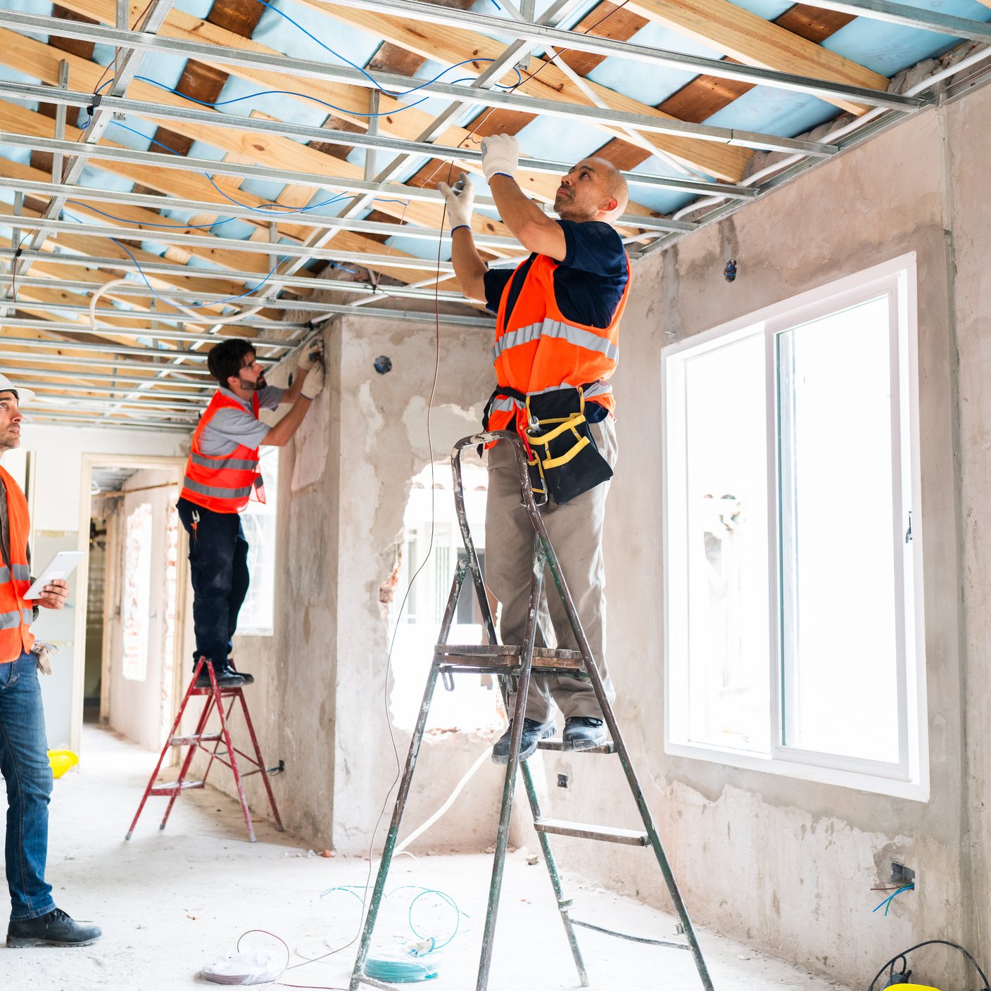 Three construction workers in safety vests and helmets work together installing ceiling framework and windows during an interior renovation project.
